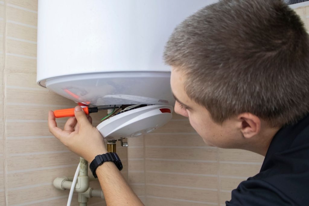 A close-up of a young male technician with short blond hair, wearing a black shirt, using a screwdriver to perform maintenance or repair on the electrical components of a white electric water heater.
