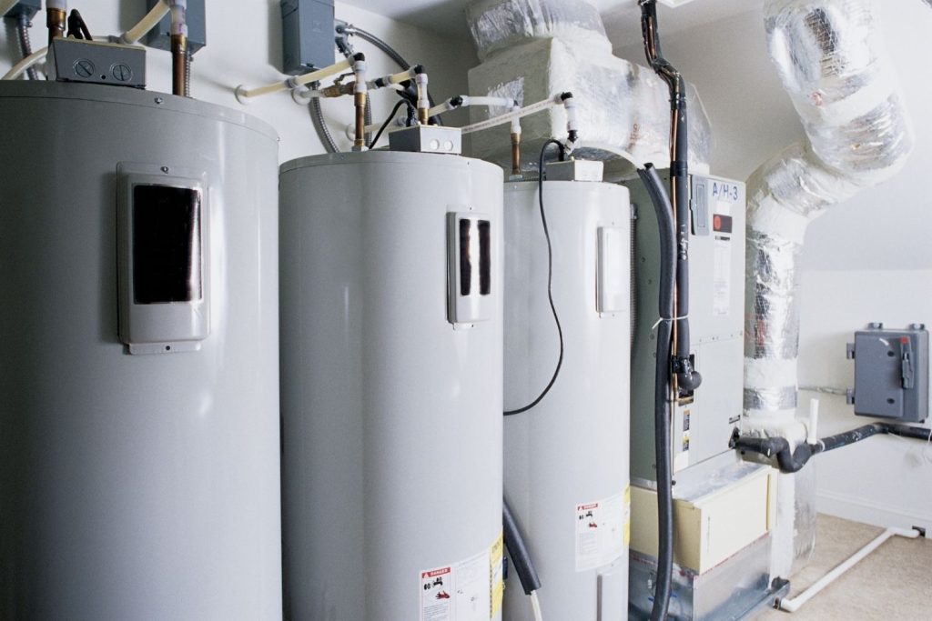 An interior view of a utility room or basement showing a row of three large, grey electric water heaters.