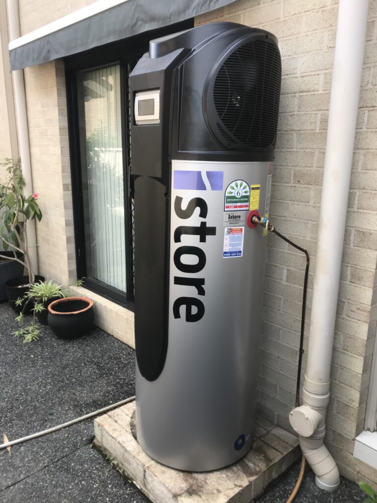 iSTORE HEAT PUMPS » iSTORE HEAT PUMPS An outdoor silver and black iStore air-to-energy heat pump system installed against white horizontal siding.