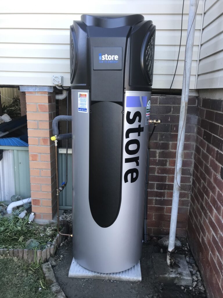 iSTORE HEAT PUMPS » iSTORE HEAT PUMPS An outdoor silver and black iStore air-to-energy heat pump system installed against white horizontal siding.
