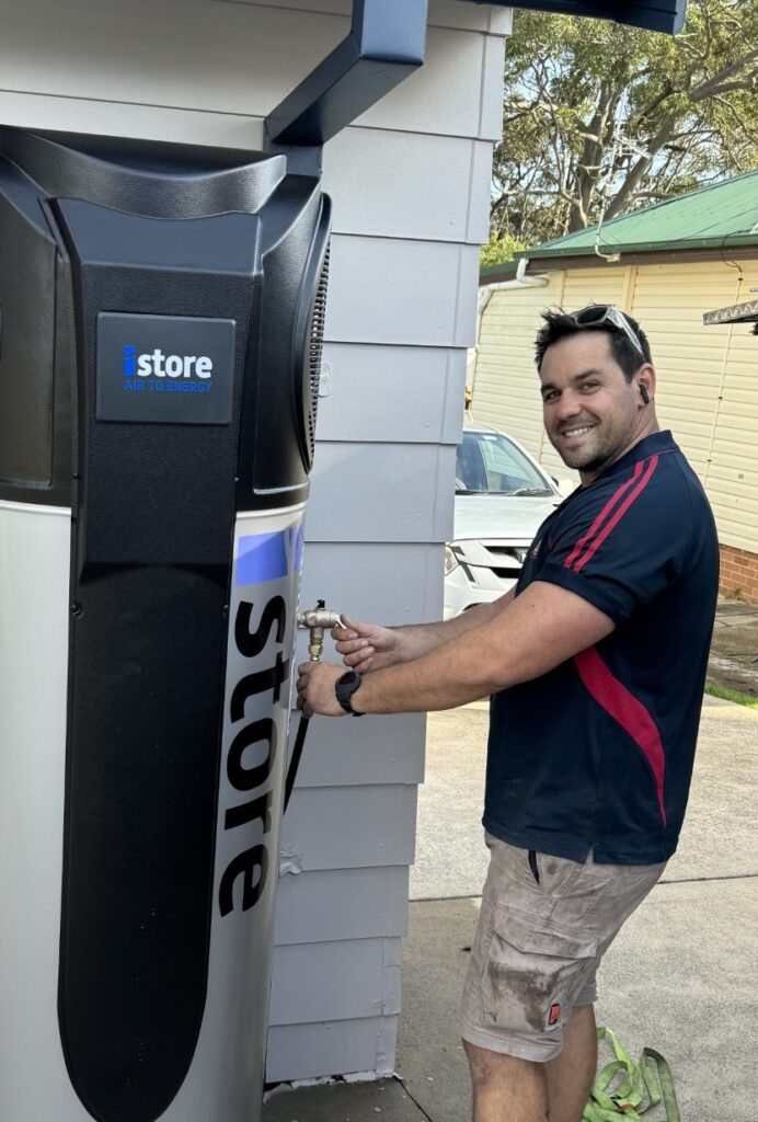 iSTORE HEAT PUMPS » iSTORE HEAT PUMPS A technician smiling while installing or servicing an iStore air-to-energy heat pump system outside a residence.