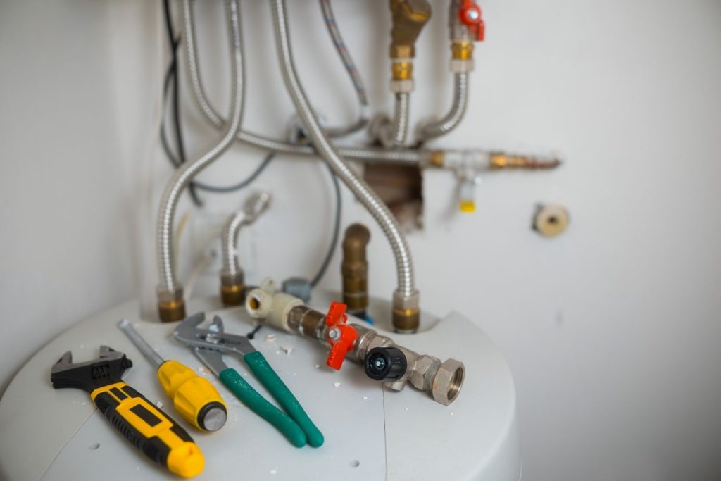 Rheem Heat Pump » Plumbing tools, including an adjustable wrench, a screwdriver, and pliers, lying on top of a water heater with flexible metal pipes in the background.