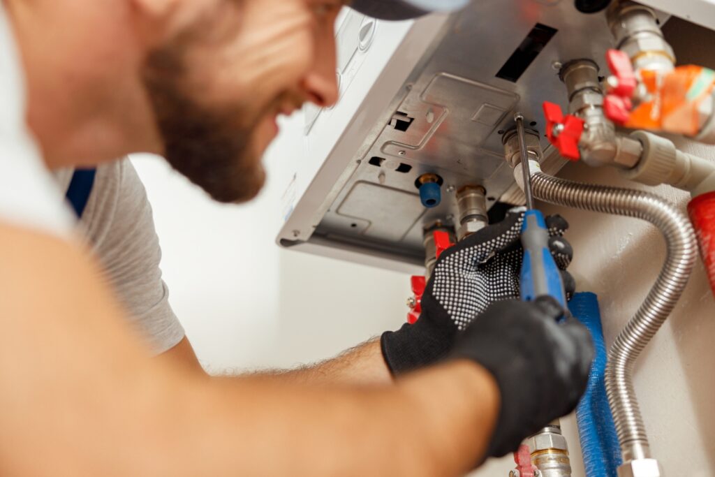 Hot Water Installation and Maintenance in Erina Heights » Hot Water A technician wearing black gloves and using a screwdriver to repair or install a wall-mounted water heater system.