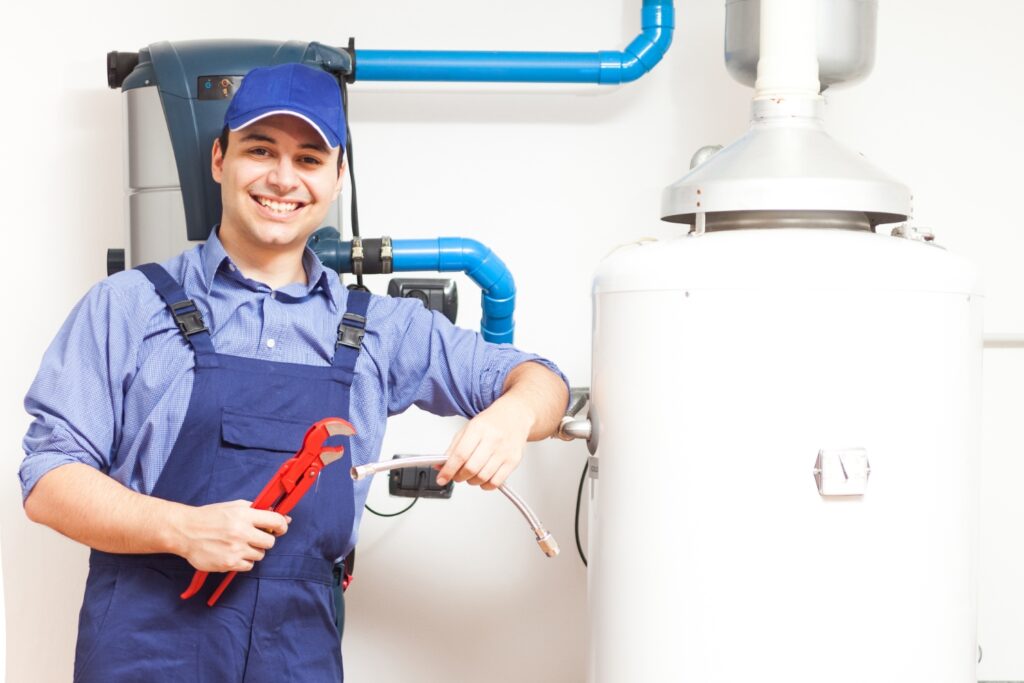 Hot Water Installation and Maintenance in Forresters Beach » Hot Water A smiling plumber in blue overalls and a cap holding a pipe wrench and a flexible hose next to a large water heater tank.