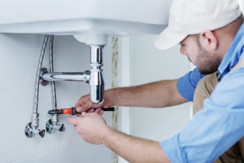 Hot Water Installation and Maintenance in Wamberal » Hot Water A plumber using a wrench to tighten or adjust the pipe fittings under a white bathroom sink.