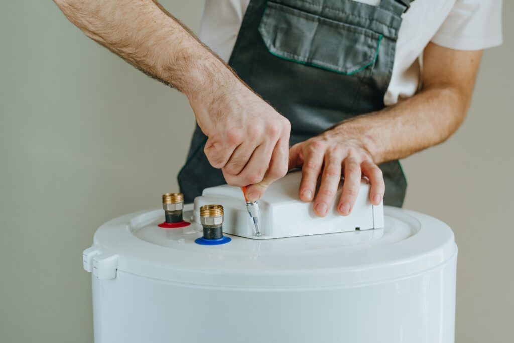 Hot Water Installation and Maintenance in Erina Heights » Hot Water A technician in grey overalls using a screwdriver to secure a white component on top of a cylindrical water heater tank.