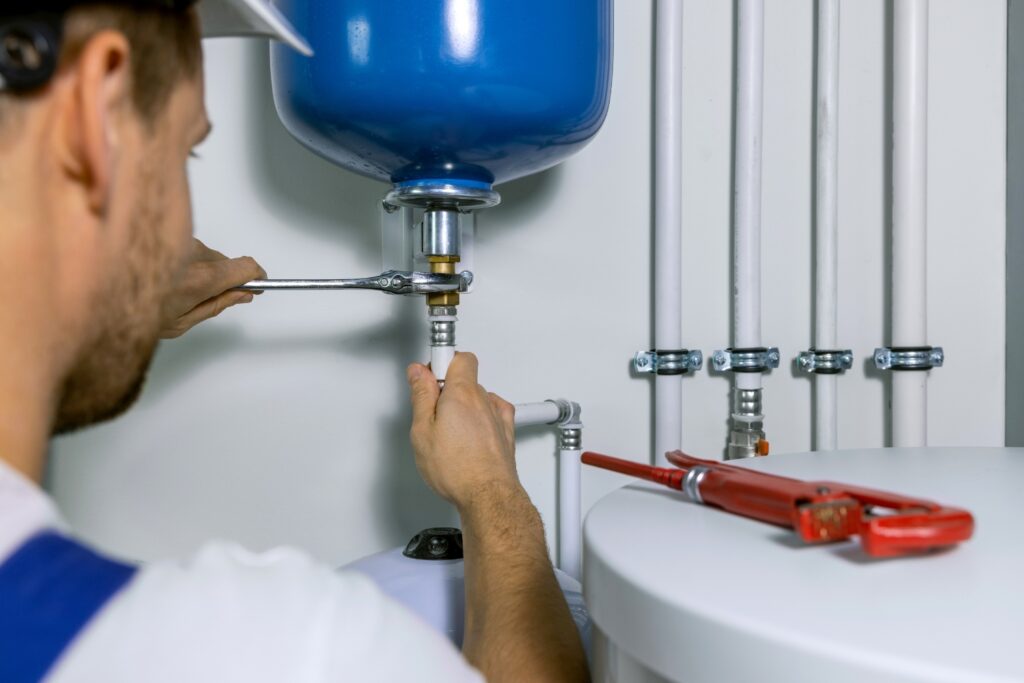 Hot Water Installation and Maintenance in Wamberal » Hot Water A plumber using a wrench to install or repair a pipe fitting connected to a blue expansion tank on a water heating system.