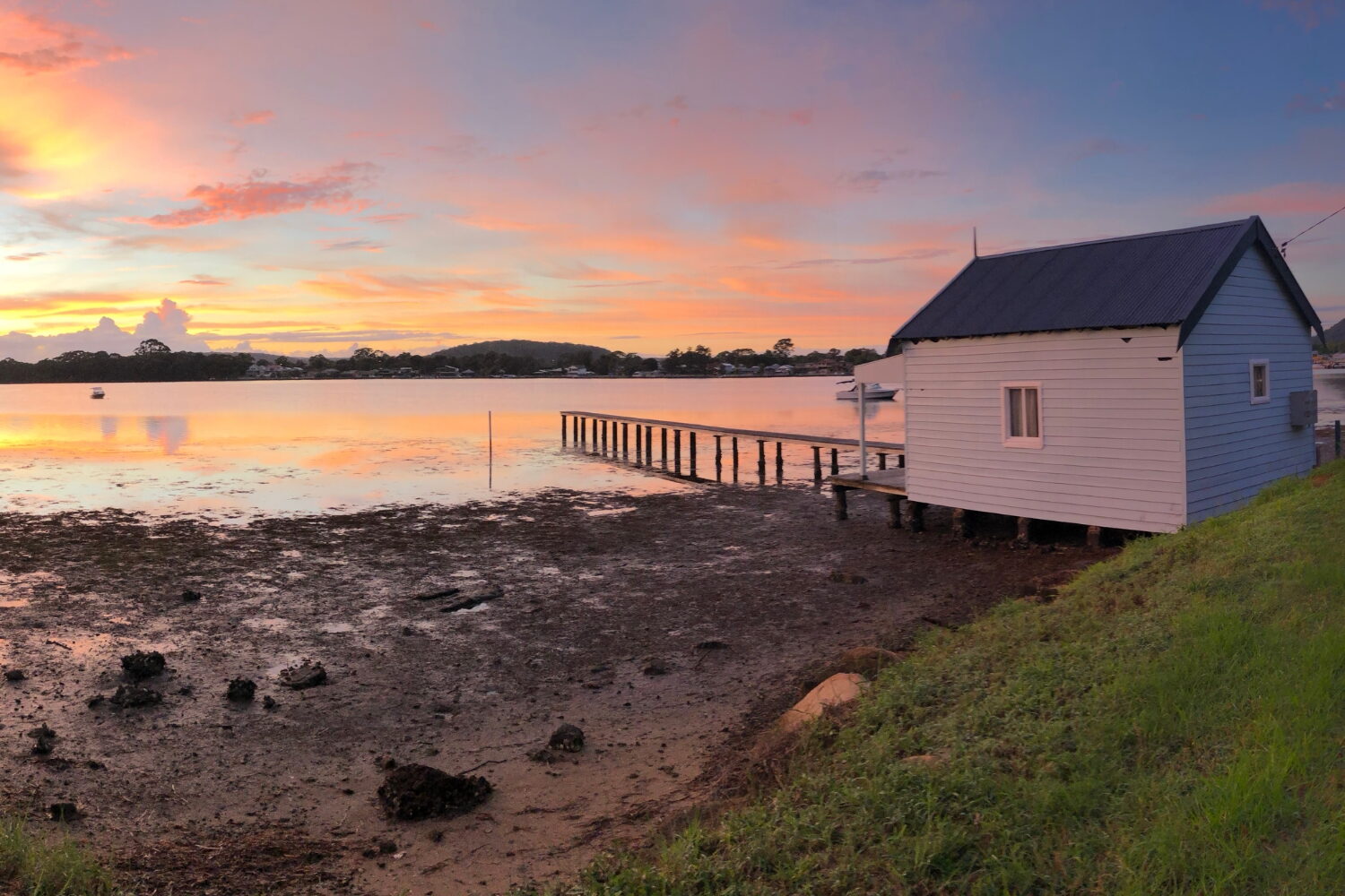 Locations » Hot Water A scenic sunset view over a calm bay during low tide, featuring a small white boatshed on stilts with a wooden jetty extending into the reflective orange and pink water.