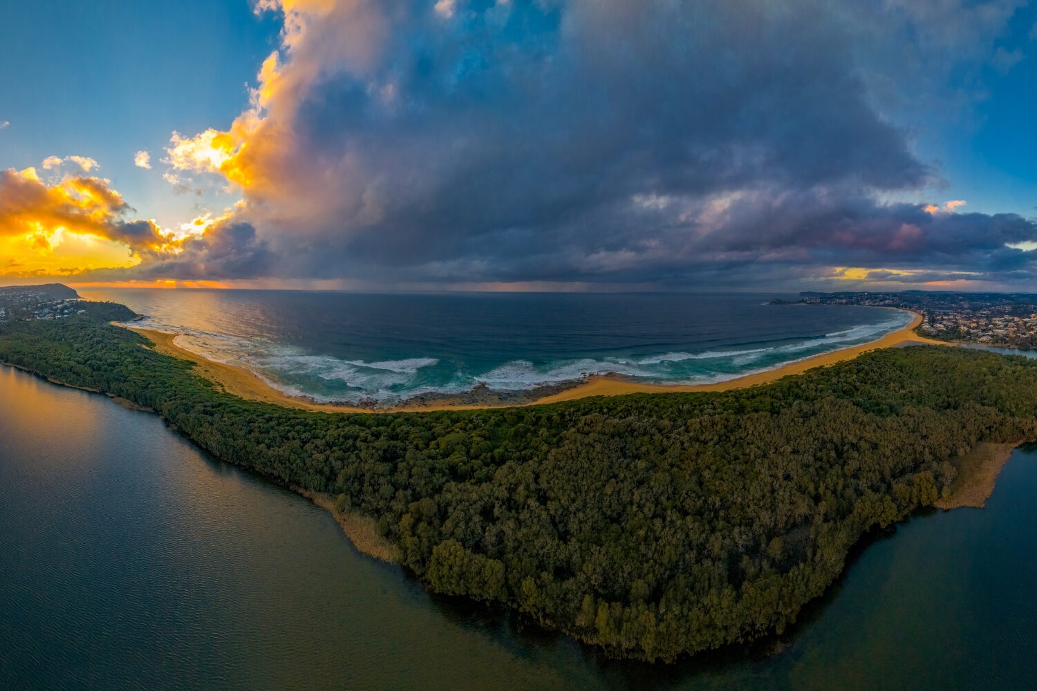 Locations » Hot Water A crescent-shaped sandy beach and ocean bay separated from an inland waterway by a thick forest, under a dramatic sunrise sky with dark storm clouds.