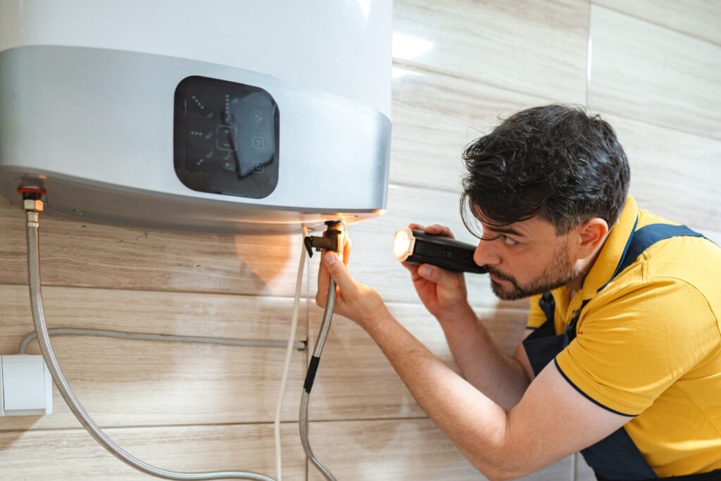 Hot Water Installation and Maintenance in Forresters Beach » Hot Water A technician in a yellow polo shirt shining a flashlight onto the connection valves underneath a modern tankless water heater.