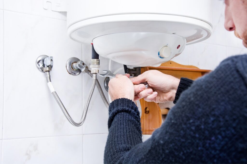 Hot Water Installation and Maintenance in Wamberal » Hot Water A technician using a small spanner wrench to adjust or repair the pipe connections underneath a white water heater unit mounted on a tiled wall.