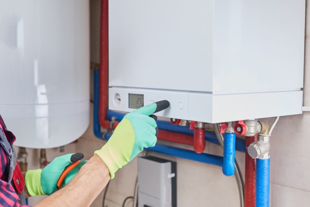 Hot Water Installation and Maintenance in Bolwarra » Hot Water A close-up of a technician wearing green and yellow work gloves adjusting a button on a modern wall-mounted boiler system next to exposed red and blue piping.