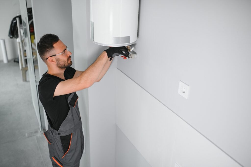 A technician in grey overalls and black gloves installing or adjusting the pipe connections underneath a wall-mounted white water heater.