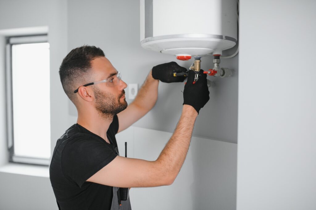 A technician wearing safety glasses and black gloves installing or repairing a wall-mounted electric water heater.