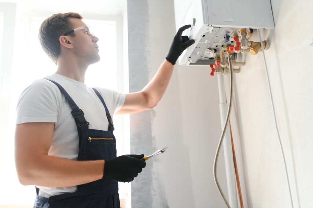 A technician wearing safety glasses and overalls holding a wrench while adjusting the settings on a wall-mounted boiler system.