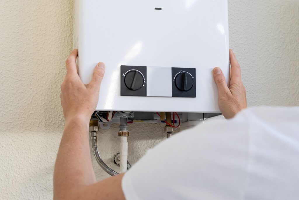 Hot Water Installation and Maintenance in Bolwarra » Hot Water A close-up view from behind a technician holding or installing a white wall-mounted water heater with two black control dials on the front panel.