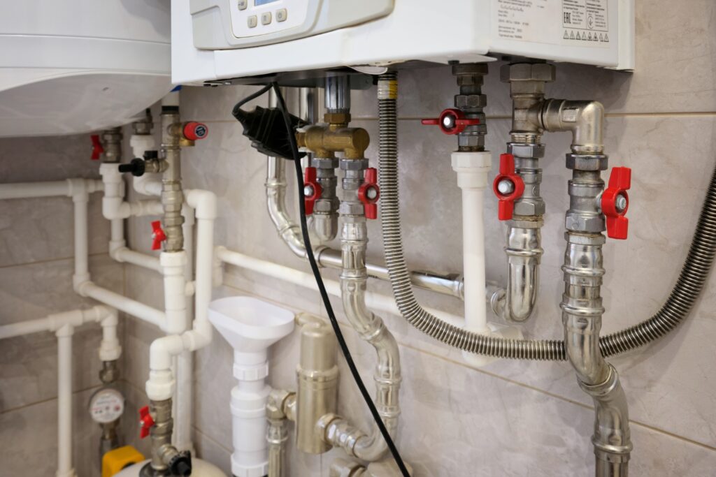 A close-up view of the complex pipe system underneath a wall-mounted boiler, featuring multiple silver metal pipes, white plastic tubes, and red shut-off valves.