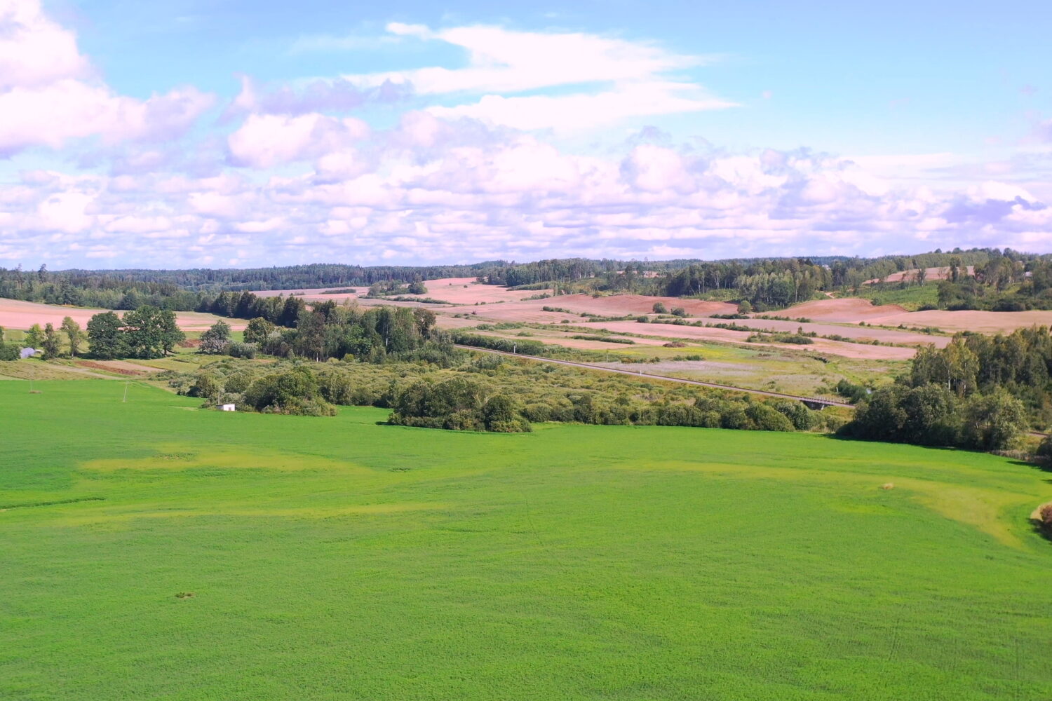 Locations » Hot Water An expansive aerial view of a green rural landscape with rolling hills, patches of trees, agricultural fields, and a cloudy sky.