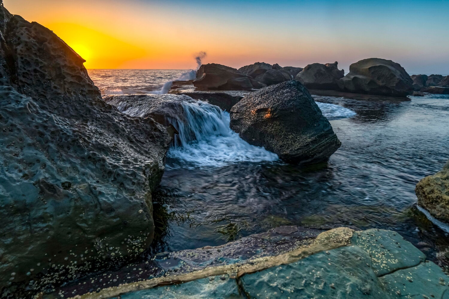 Locations » Hot Water A scenic sunrise over a rocky coastline with ocean water cascading between large boulders.
