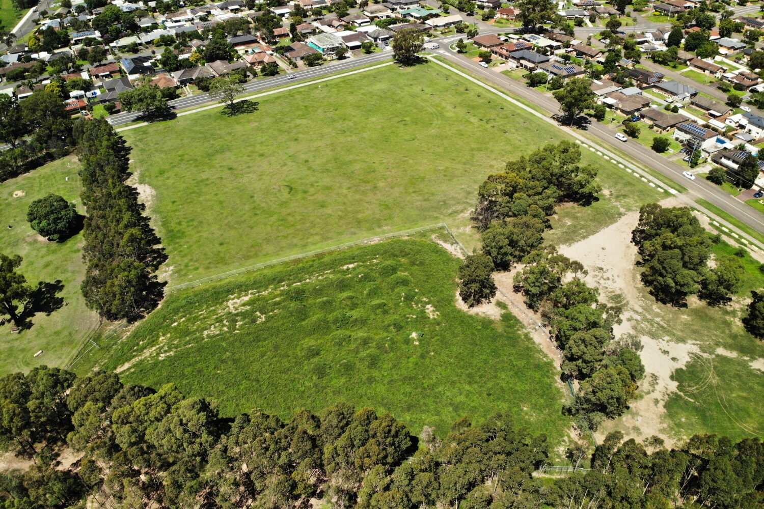 A large, green grassy field surrounded by trees, bordered by a suburban residential neighborhood with houses and streets.