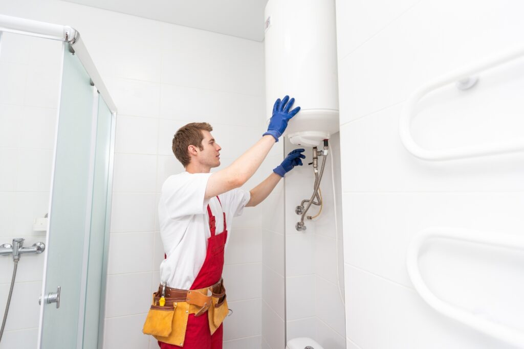 Hot Water Installation and Maintenance in East Maitland » Hot Water A plumber wearing a white t-shirt, red overalls, a tool belt, and blue gloves works on a wall-mounted cylindrical water heater inside a bright white bathroom.