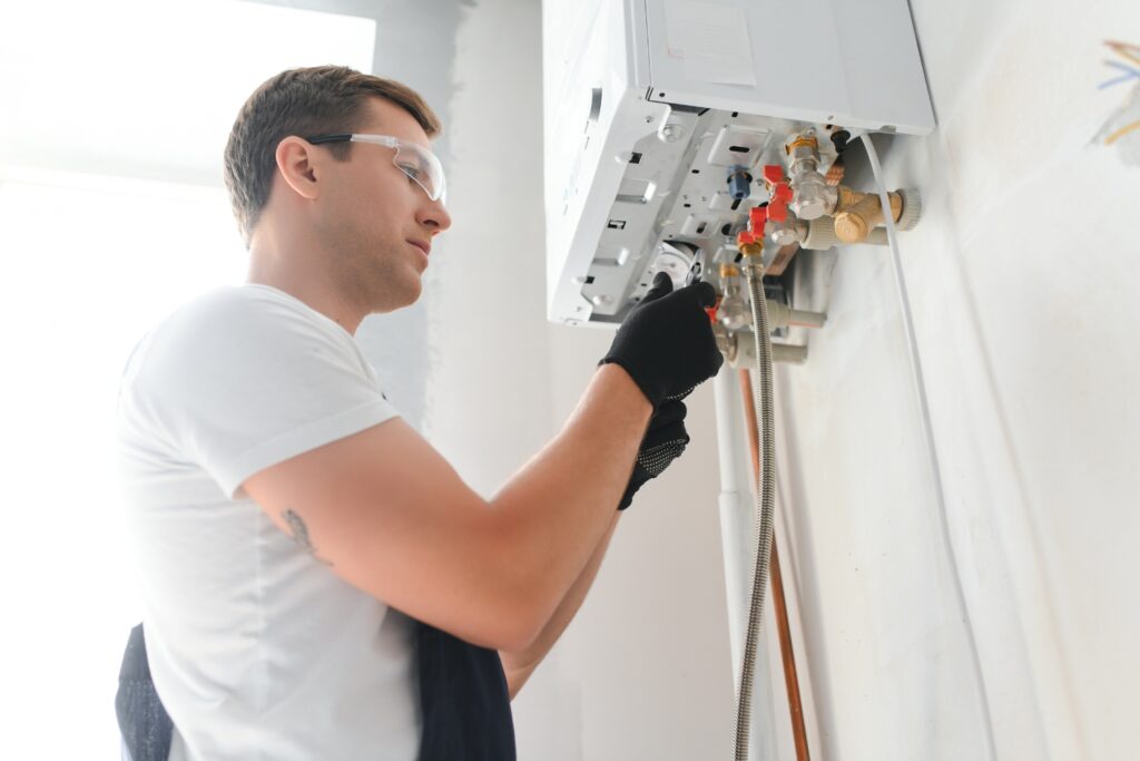 A technician wearing safety glasses and grey overalls installing or repairing the valve connections beneath a white, wall-mounted water heater.