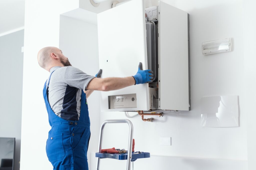 A technician in blue overalls removing the front cover panel of a wall-mounted water boiler.