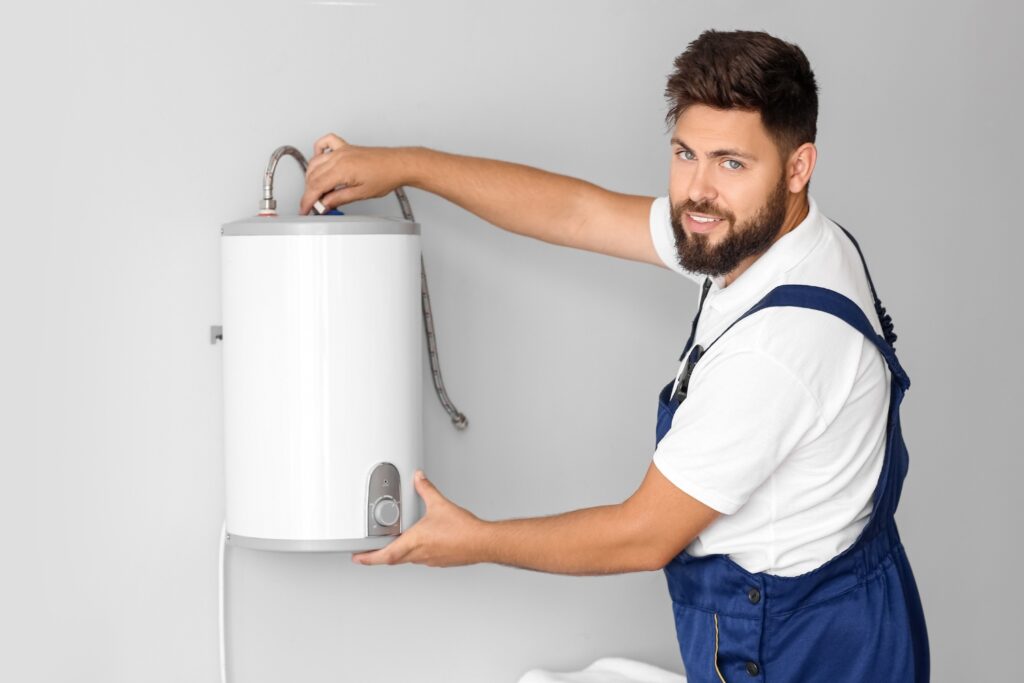 Hot Water Installation and Maintenance in Metford » Hot Water A smiling technician with a beard, wearing blue overalls, adjusts a small white cylindrical water heater mounted on a grey wall.