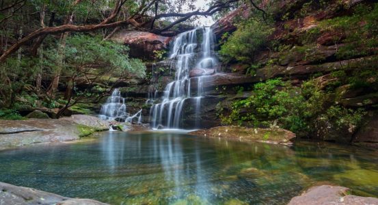 A long-exposure view of a multi-tiered waterfall cascading down a red rock face into a clear pool of Kariong