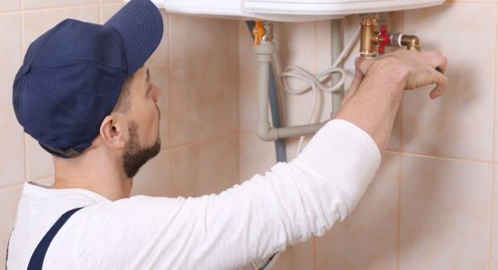 Hot Water Installation and Maintenance Matcham A technician wearing a blue cap and white long-sleeved shirt working on the pipe fittings beneath a white wall-mounted hot water heater unit.