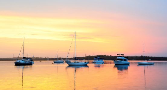 Hot Water Mannering Park Several sailboats and motorboats anchored on a glassy, still lake during a pastel sunset.