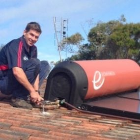 heater A technician in a dark blue polo with red stripes smiling while using a wrench to work on the plumbing