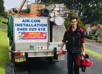 img-about A technician in a black and red jacket standing next to the rear of a Hogan Hot Water & Air Conditioning service truck