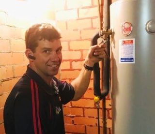 img1-a A technician in a dark blue polo shirt smiling while adjusting a valve on a tall, silver Rheem hot water heater