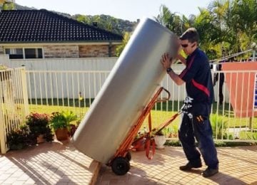 About Us » Hot water systems A technician in a dark blue uniform uses an orange hand truck to carefully move a large, silver hot water tank