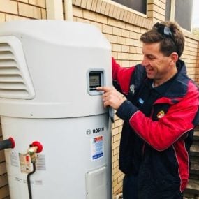 About Us » Hot water systems A technician in a red and dark blue jacket smiling while operating the digital control panel of a large, white Bosch heat pump hot water system