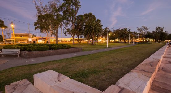 A tranquil park scene at dusk featuring a paved walkway lined with palm trees and streetlights